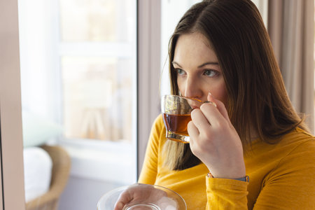 Thoughtful caucasian woman drinking tea by window in living room at home, copy space. Expression,...