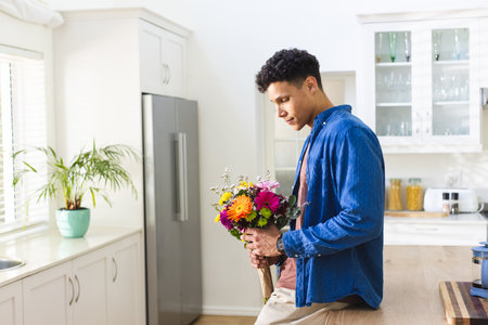 Happy biracial man holding flowers in kitchen at home, copy space. Expression, plant, gift and domestic life unaltered.の写真素材
