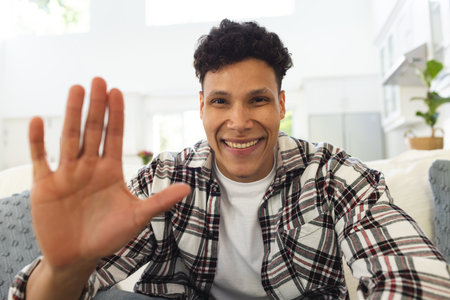 Portrait of happy biracial man sitting on couch and waving with hand at home, copy space. Expression, leisure, gesticulation and domestic life unaltered.の写真素材