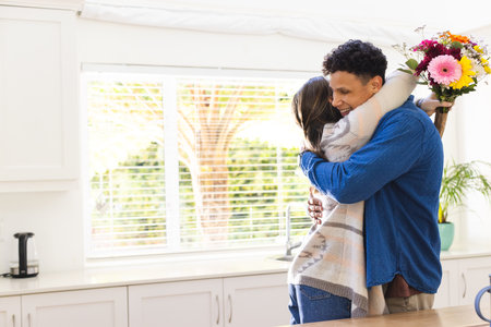 Happy diverse couple holding flowers and embracing in kitchen at home, copy space. Expression, love, togetherness, plant, gift and domestic life unaltered.の写真素材