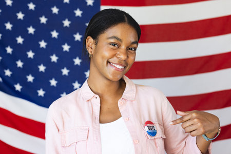 Young biracial woman poses proudly with her vote badge in front of an American flag, promoting civic...