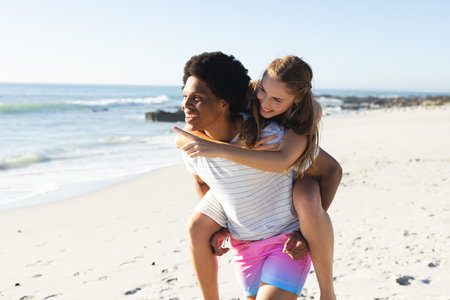Young African American man gives piggyback ride to Caucasian woman on beach. Their joyful moment captures the essence of carefree summer days outdoors.の写真素材