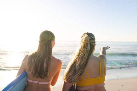 Two young Caucasian women gaze at the ocean, with copy space. They are ready for a surf session at...