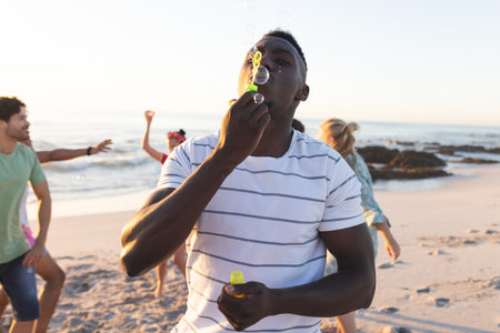Young African American man blows bubbles on the beach. Friends enjoy a playful moment together in an outdoor setting.の写真素材