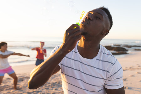 Young African American man blows bubbles at the beach. Friends enjoy a playful moment in the outdoor...