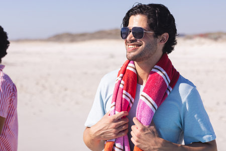 Young biracial man enjoys a sunny beach day, with copy space. He's outdoors, smiling with sunglasses and a towel around his neck.の写真素材