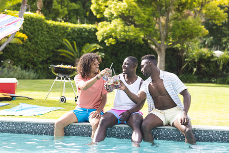 Young African American man and biracial man enjoy drinks by the pool with a friend. Casual summer...