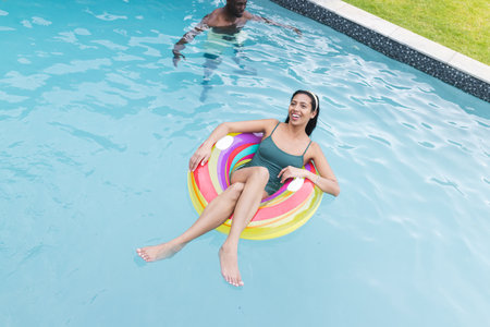 A young biracial woman lounges on a colorful pool float, smiling. In the background, a young African...