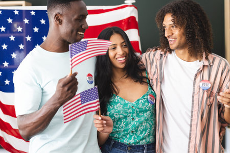 African American man and biracial couple hold American flags, all wearing I Voted stickers. They stand before a large American flag, smiling, having participated in an election.の写真素材