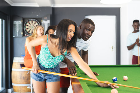 Biracial woman in a green top plays pool, guided by a young African American man. Friends cheer in...