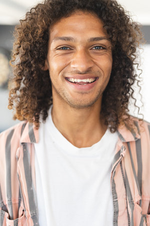 Young biracial man with curly brown hair smiles warmly. He wears a white t-shirt with a striped pink overshirt, radiating a casual, friendly vibe.の写真素材