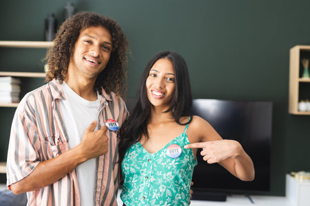 Biracial couple proudly displaying I Voted stickers, standing in a home setting. Young man with...