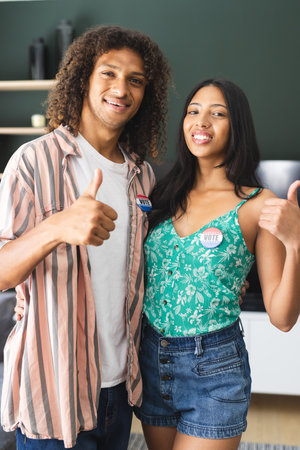 Biracial couple with I Voted stickers give thumbs up, smiling confidently. They stand in a home...