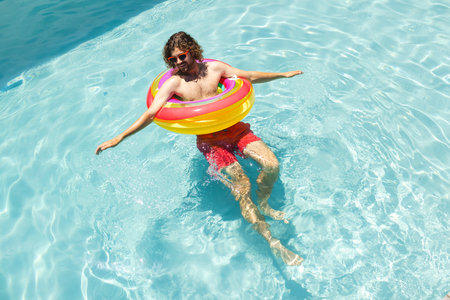 Young Caucasian man enjoys a sunny day in the pool with a colorful float ring. Wearing sunglasses...