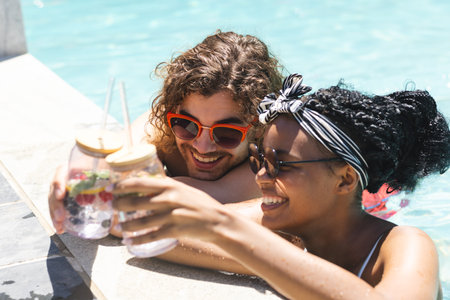 Diverse couple enjoying drinks by the pool, the woman's hair adorned with a headband. Their cheerful mood and summer attire suggest a relaxing vacation vibe.の写真素材