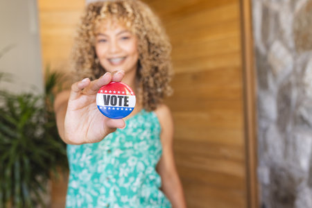 Young biracial woman holds a 'vote' badge towards the camera, with copy space. Her cheerful expression promotes the importance of participating in elections.の写真素材