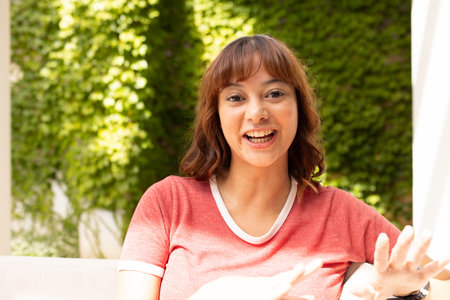 Young biracial woman smiles brightly outdoors on video call. Her cheerful expression adds a warm, inviting atmosphere to the sunny day.の写真素材