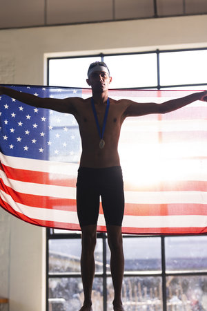 Proud young biracial male athlete swimmer displays the American flag, with copy space. He celebrates a sports victory with a medal, symbolizing achievement and patriotism.の写真素材