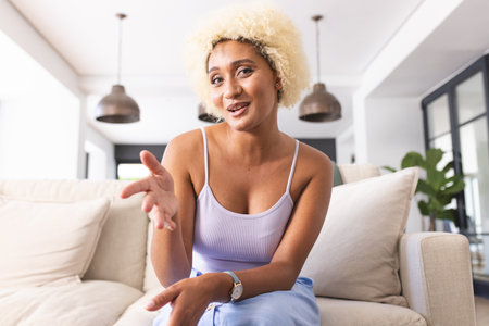 Young biracial woman with curly blonde hair extends her hand in a welcoming gesture on video call....