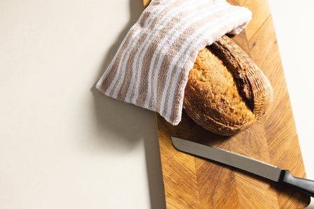 Bread fresh from the oven on a wooden board, partly hidden by a striped towel. A sharp knife lies...
