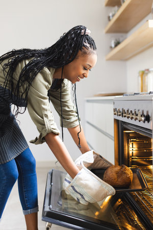 Black woman, both student and pro, with oven mitts, removes bread.. She has long braided hair, wearing casual shirt and jeans, unalteredの写真素材