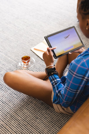 African American young woman sitting, holding tablet showing travel booking. Wearing blue plaid shirt, she has dark hair, focused on screen, unalteredの写真素材