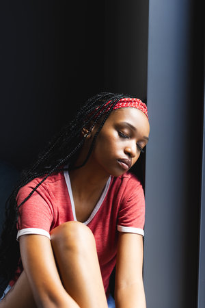 A young African American woman sitting by window, looking thoughtful. She has long braided hair, red...