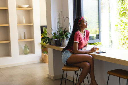 African American young woman sitting, looking out window. Wearing red shirt and blue shorts, she has dark hair and is at home, unalteredの写真素材