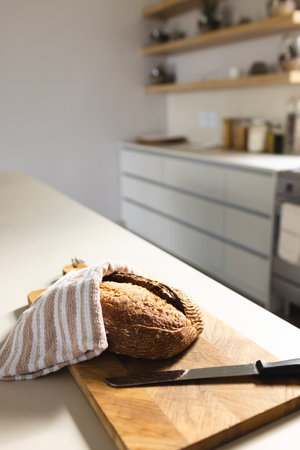 Freshly baked bread rests on wooden cutting board, covered with a striped towel. Sunlight fills...