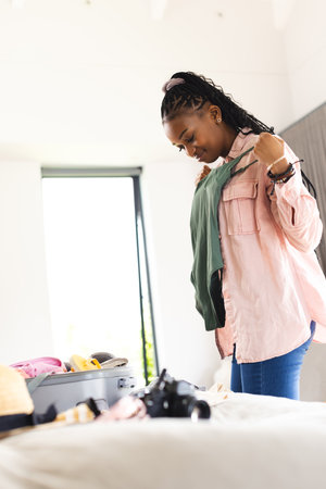 African American young woman packing clothes into suitcase. She has dark braided hair, wearing a pink shirt and jeans, unalteredの写真素材