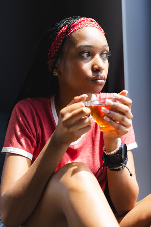 African American young woman holding cup of tea, looking thoughtful. She has dark eyes, braided hair with a red headband, and wears a red shirt, unalteredの写真素材