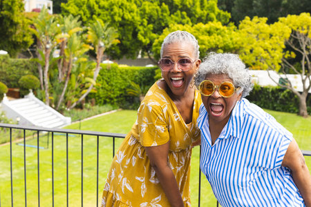 Senior African American woman and senior biracial woman share a joyful moment outdoors. Both are wearing glasses and vibrant clothing, exuding happiness and vitality in a garden setting.の写真素材