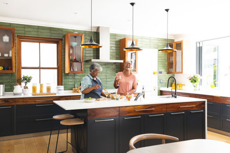Senior African American woman and senior biracial woman are preparing food in a kitchen. They share a joyful moment while cooking together, surrounded by a modern interior.の写真素材