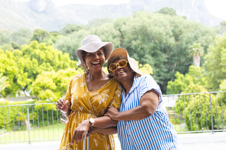Senior African American woman and senior biracial woman share a joyful embrace outdoors on vacation. Both are stylish in summer dresses and wide-brimmed hats, with a backdrop of lush greenery and mountains.の写真素材
