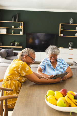 Senior African American woman and senior biracial woman share a tablet, smiling in a modern kitchen at home. Dressed in casual attire, they enjoy a moment of connection amidst vibrant home decor.の写真素材