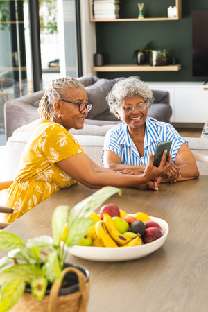Senior African American woman and senior biracial woman share a moment over a smartphone at home. They are seated at a table with a bowl of fruit, radiating joy in a cozy home setting.の写真素材