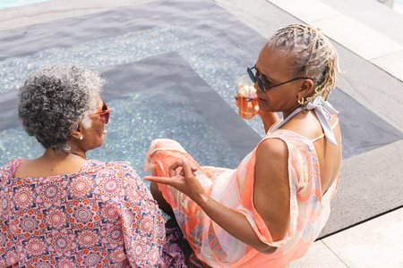 Senior African American woman and senior biracial woman enjoy a poolside chat with copy space. Both are dressed in vibrant summer attire, reflecting a leisurely day outdoors.の写真素材