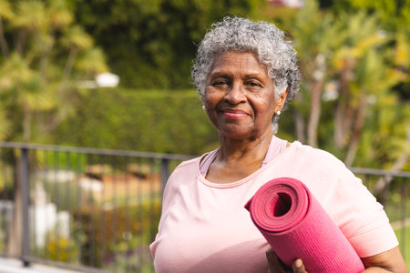 Senior biracial woman with gray hair holds a pink yoga mat outdoors. She smiles gently, suggesting a healthy lifestyle and the importance of fitness at any age.の写真素材