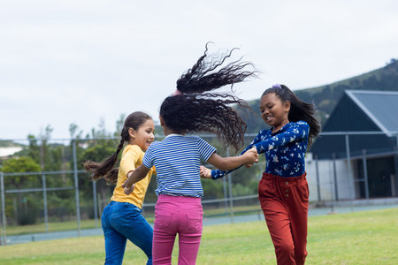 Three girls are joyfully holding hands and spinning in a grassy field in school. Their dynamic movement suggests a carefree moment of childhood play outdoors.の写真素材