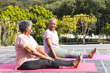 Senior African American woman and senior biracial woman are sitting on yoga mats outdoors. They are smiling and engaging in a stretching exercise, surrounded by greenery in a tranquil setting.の写真素材