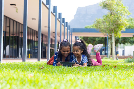 Two biracial girls are engrossed in a tablet outdoors at school, lying on the grass. They enjoy a sunny day at school with a mountain in the background.の写真素材