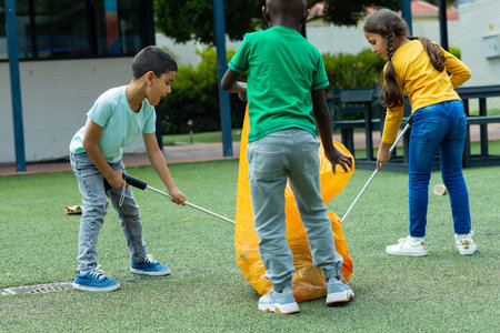 Three children are collecting trash in school. A biracial girl in a yellow shirt assists an African...