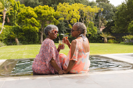Senior African American woman and senior biracial woman enjoy drinks by the pool. They are dressed in summer attire, sharing a moment of relaxation in an outdoor setting.の写真素材