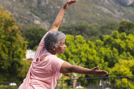 Senior biracial woman with gray hair practices yoga outdoors. She wears a pink top, demonstrating a pose that reflects tranquility against a backdrop of greenery.の写真素材
