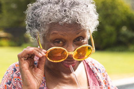 Senior biracial woman with curly gray hair lowers her oversized yellow sunglasses. She is smiling...