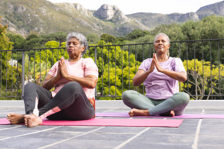 Senior African American woman and senior biracial woman are practicing yoga outdoors. They are seated on yoga mats, performing a pose with a serene mountain backdrop.の写真素材