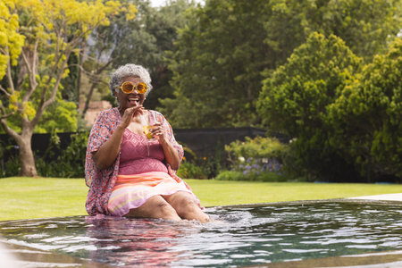 Senior biracial woman enjoys a drink by the poolside, her feet dipped in water, with copy space. She...