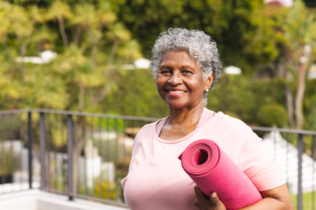 Senior biracial woman with gray hair holds a pink yoga mat outdoors. She smiles warmly, showcasing an active lifestyle and well-being in her golden years.の写真素材