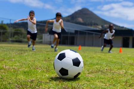 A soccer ball in focus with three children running behind it in school. Biracial and African American boys are playing soccer outdoors on a sunny day.の写真素材