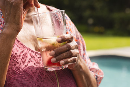 Senior biracial woman enjoys a refreshing drink by the pool, her hand delicately holding a glass. Wearing a pink top, she embodies relaxation and leisure on a sunny day.の写真素材